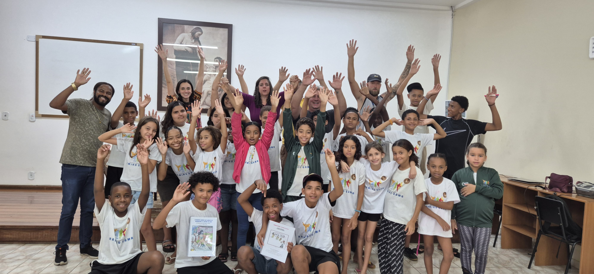 A large group of children, project leaders, and volunteers from Moleques do Bem raising their hands and smiling together in celebration.
